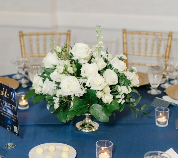 A low white rose and greenery centrepiece on navy linen with gold chairs and votives