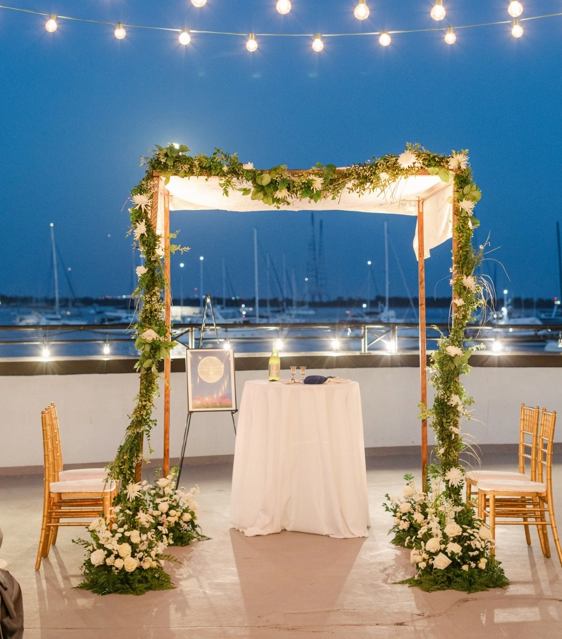 A waterfront chuppah lit with string lights at dusk beside the marina