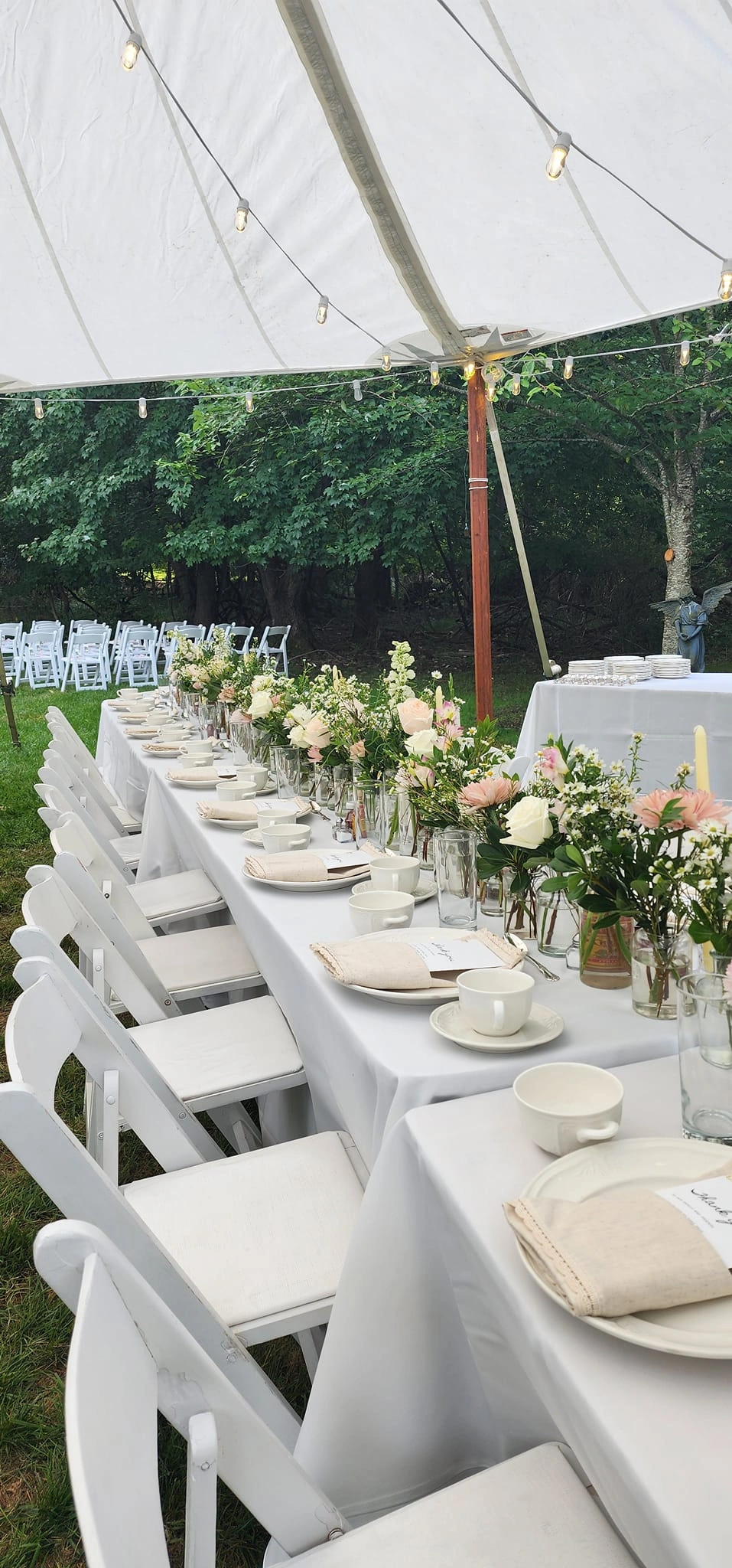 A long garden table laid with peach and cream arrangements and white chairs beneath the trees