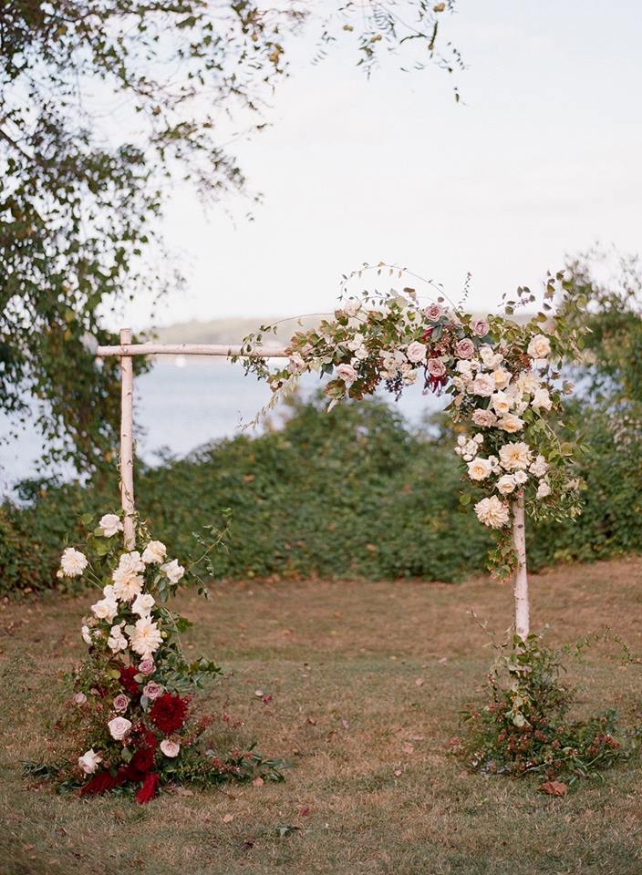 A birch arbour arch dressed with cream and mauve roses and burgundy dahlias, overlooking the water in autumn