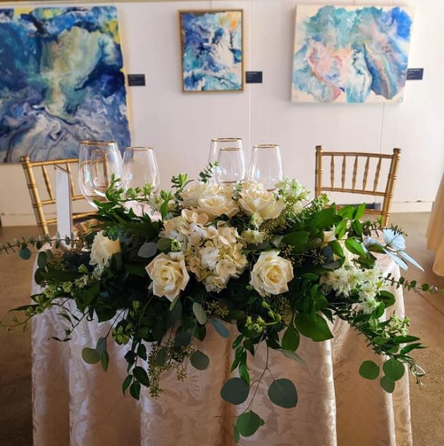 A sweetheart table topped with a white rose and greenery garland in a gallery hung with blue paintings
